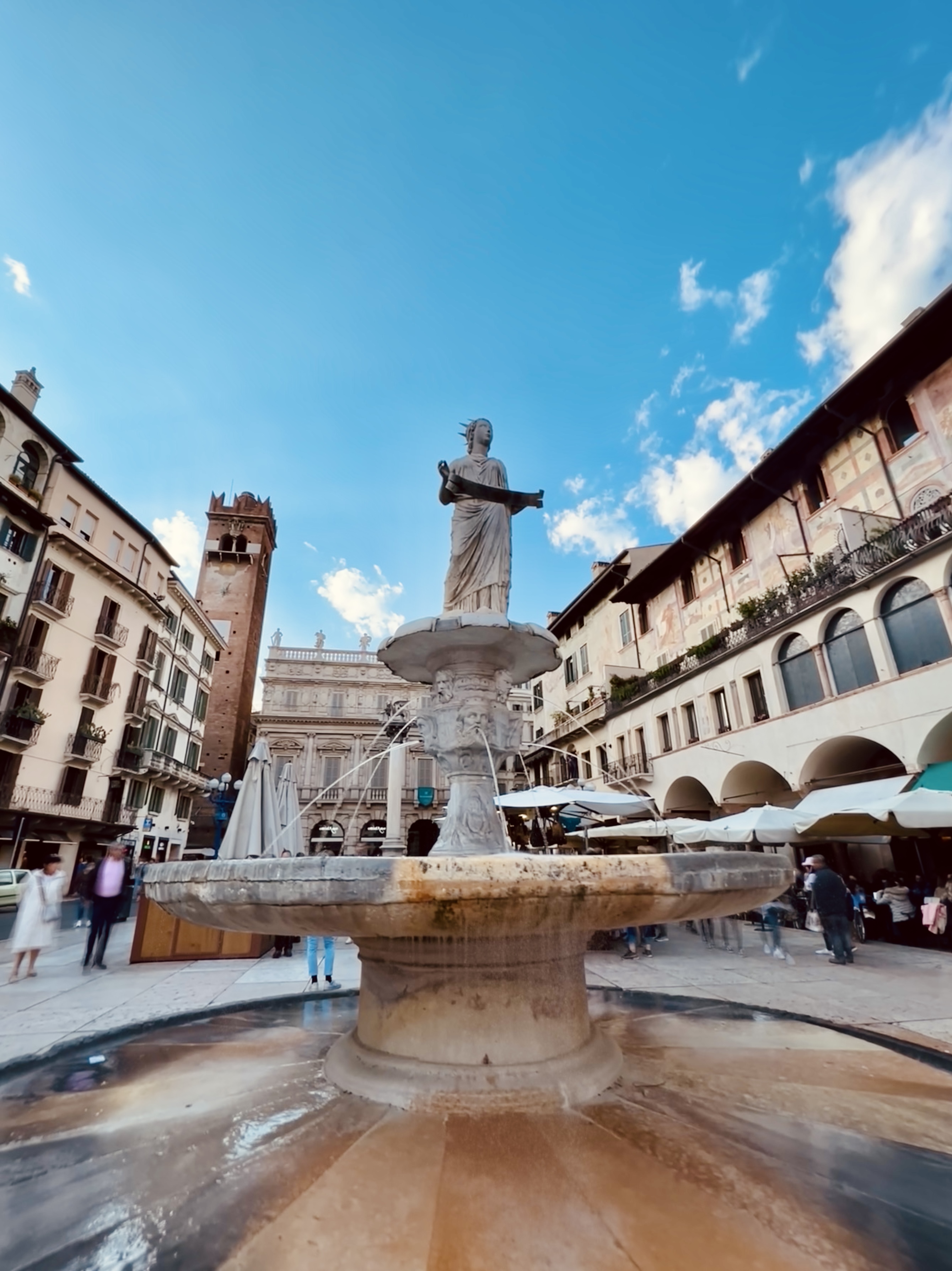 Ancient Roman fountain with a robed woman surrounded by buildings with frescoes and a brick tower in Verona, Northern Italy