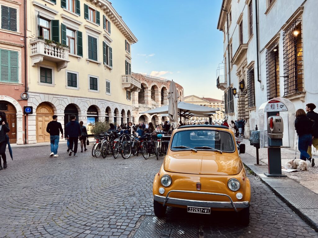 Small, yellow car on cobblestone streets surrounded by red, yellow, and white buildings in front of the Verona Arena in Verona, Northern Italy