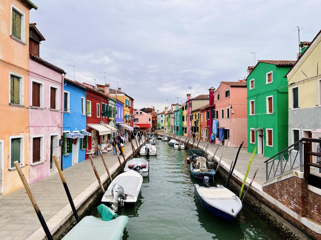 A jade green canal lined with boats surrounded by orange, pink, blue, purple, red, and green buildings in Burano, Northern Italy