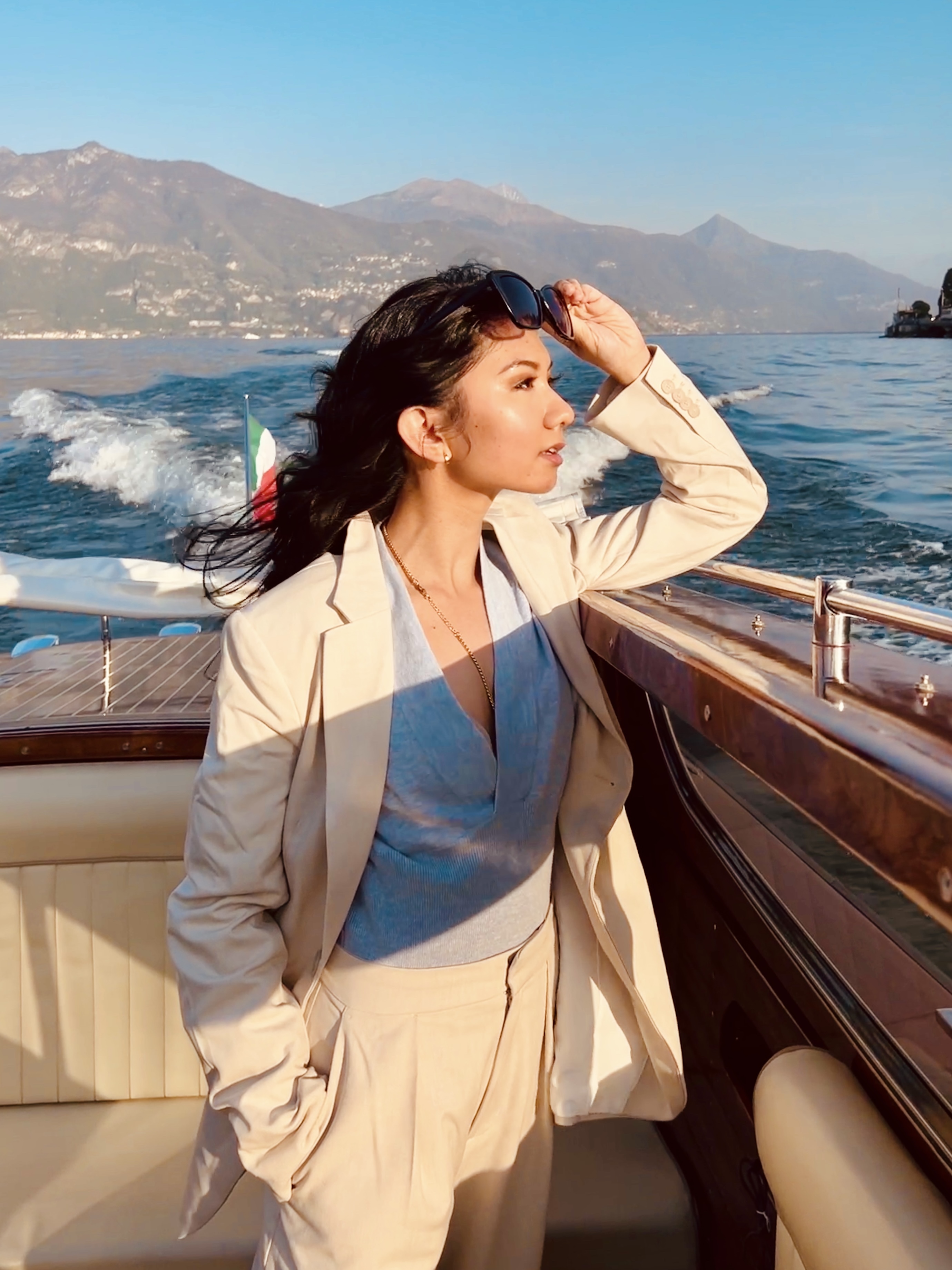 Woman in blue shirt, white blazer, and white pants on a Venetian taxi boat looks out onto the waters of Lake Como