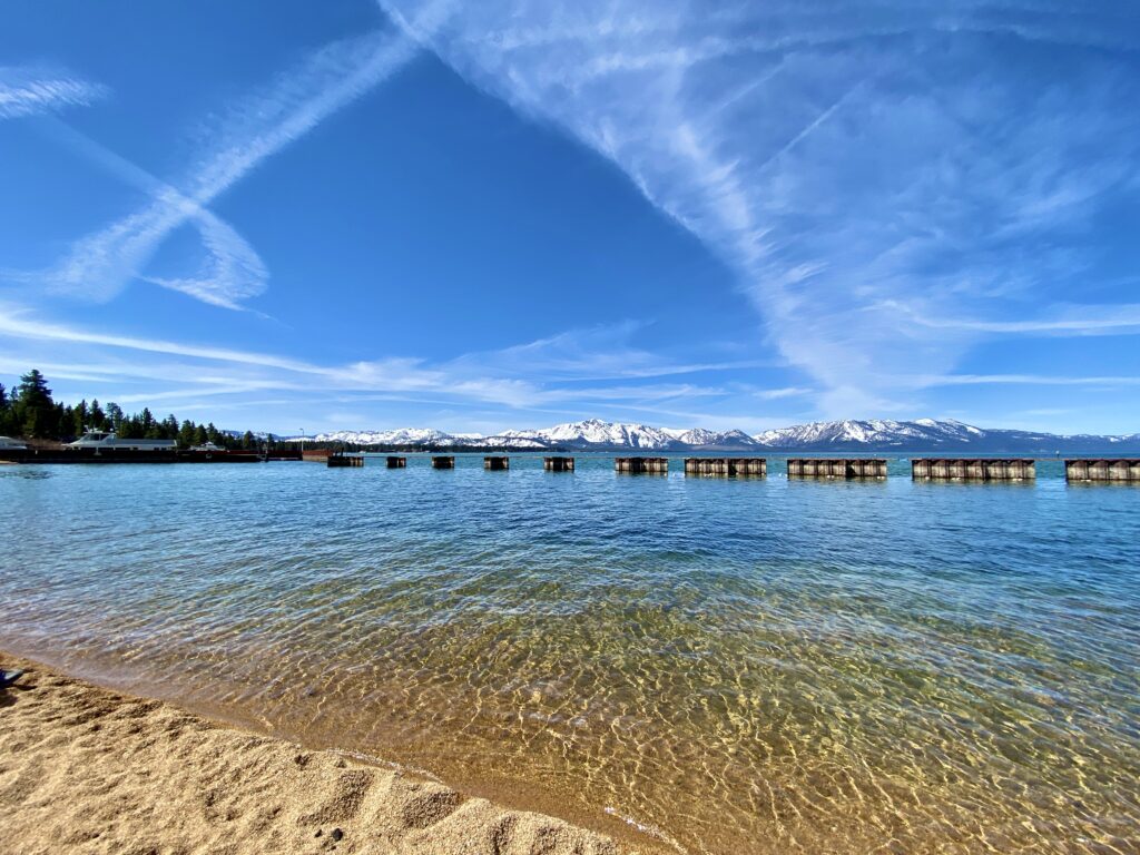 Lakeside Beach at Lake Tahoe featuring yellow and blue lake waters, a wooden pier, and snow-capped mountains