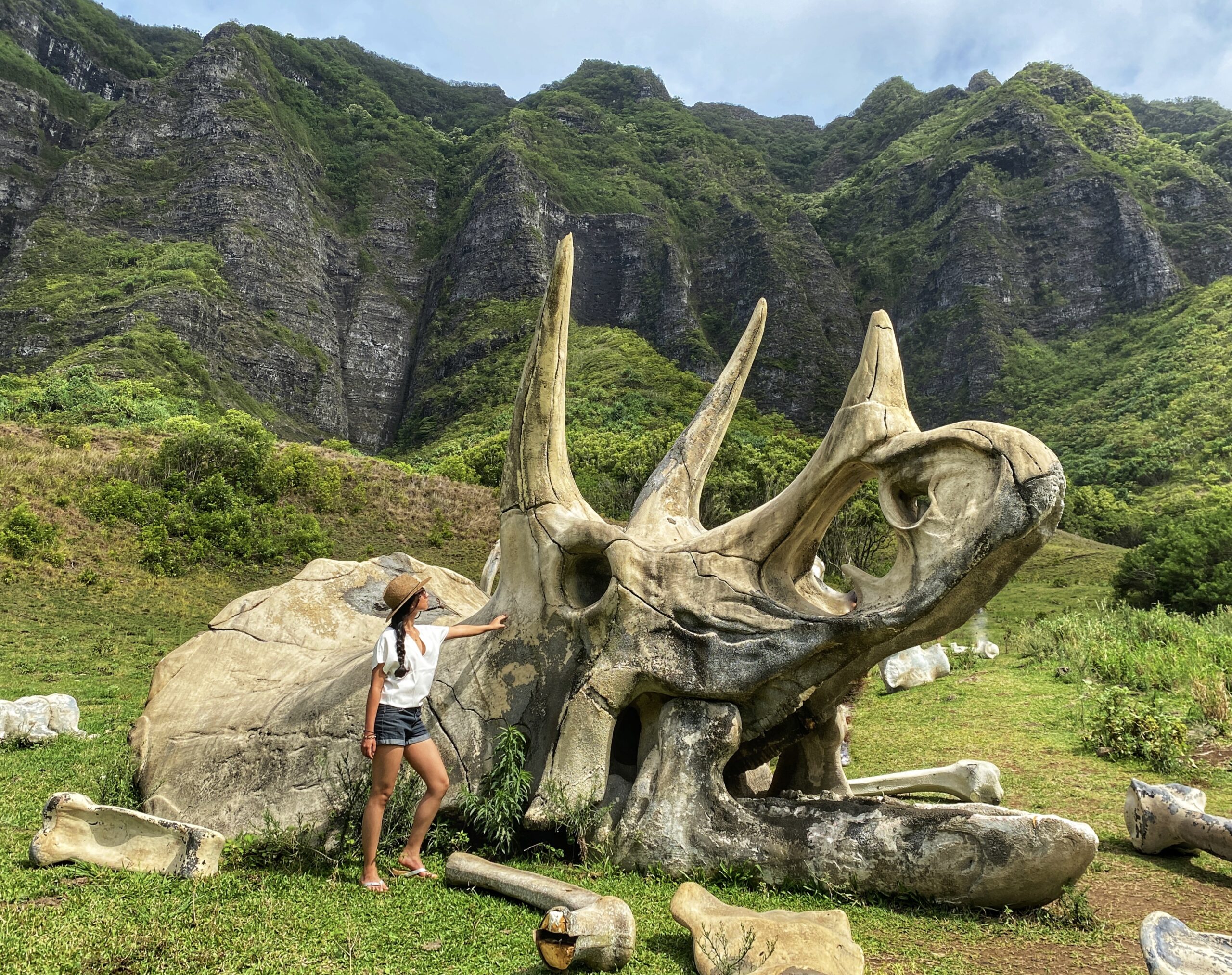 Woman in a hat, white shirt, and shorts standing in front of a triceratops fossil movie set piece from kong skull island